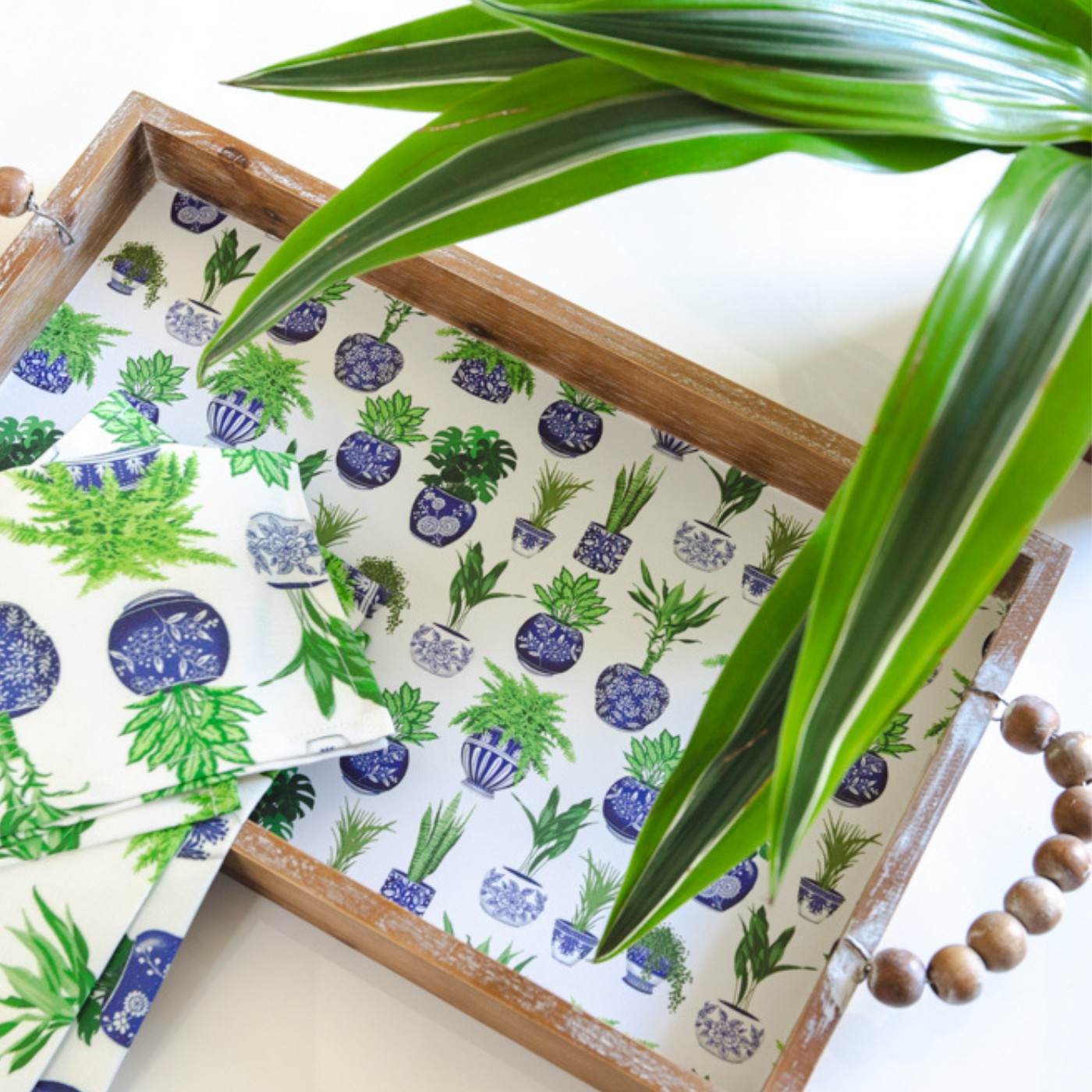A wooden tray with curved handles comprised of small wooden beads sits on a white table with a plant with long extended leaves. Some of the plant fronds are hanging over the tray. The surface of the tray is covered in a design with a white background and rows of small green plants in blue pots. Napkins in the same design are laying at the bottom left of the tray.
