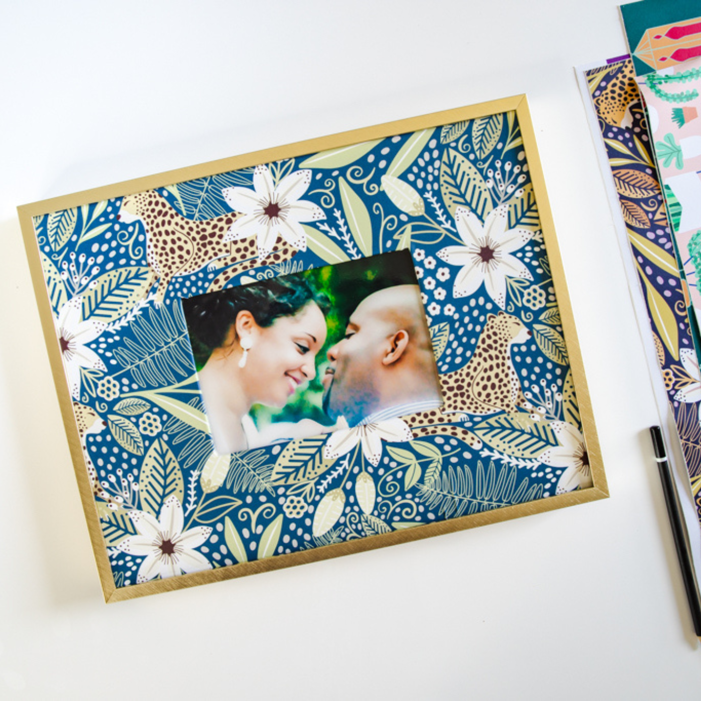 A photo of two people standing with their faces close together sits in a photo frame with a peel-and-stick wallpaper border. The print on the wallpaper features leopards and bright blue flowers as well as white flowers. The photo frame is sitting on a white table next to a black pen and several pieces of brightly colored paper.