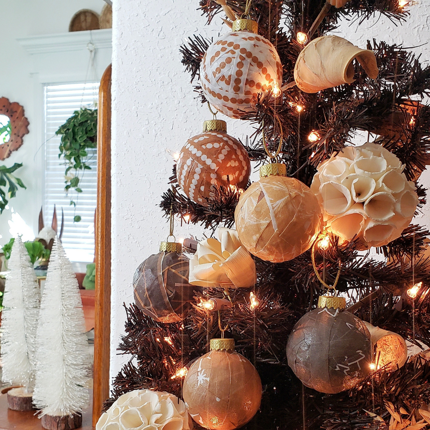 Round glass ornaments wrapped in strips of varying shades of brown fabric, some with white dots, some with white snowflakes, adorn a green fir tree with small warm golden lights.
