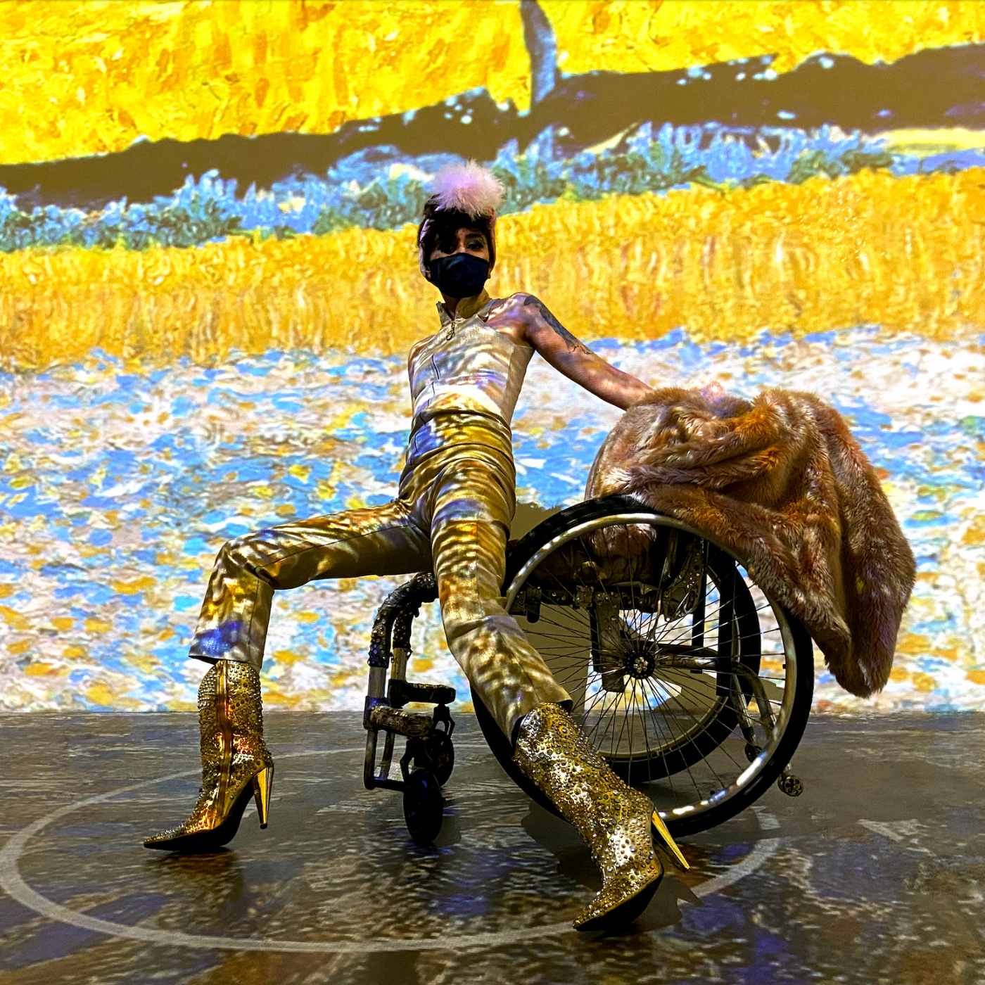 Pansy St. Battie is on stage wearing gold-toned clothing, holding on to a wheelchair.