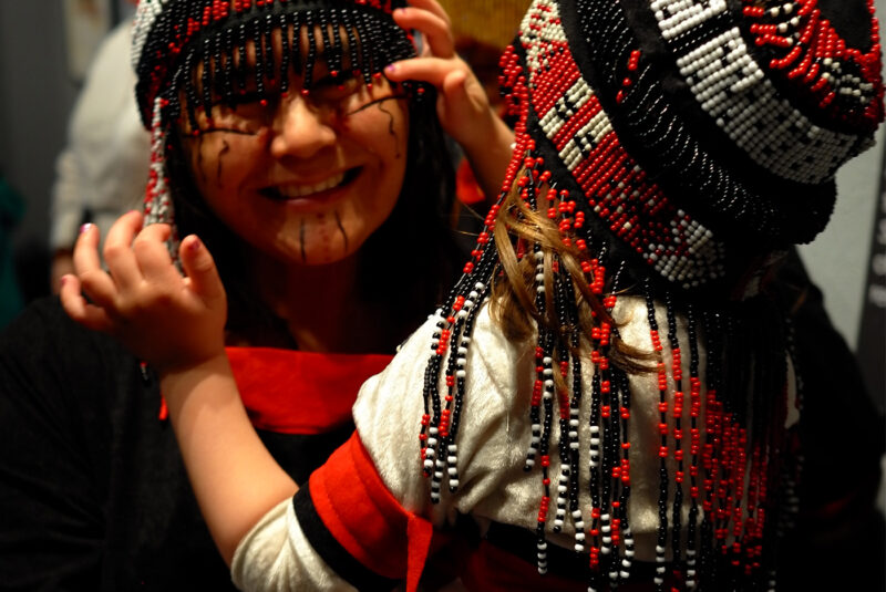 A smiling adult holds a small child while both wear beaded headpieces and enjoy each other’s company at the Alutiiq Museum in Alaska. The Alutiiq Museum or Alutiiq Museum and Archaeological Repository is a non-profit museum and cultural center dedicated to preserving and sharing the cultural traditions of the Koniag Alutiiq branch of Sugpiaq ~ Alutiiq of the Alaska Native people.