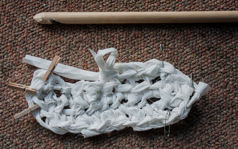 The beginnings of the rag rug with chain stitches in white fabric running through the center, and single crochet (US single crochet) extending at the top and bottom. Safety pins are holding the edge stitches together on the right and small wooden clothespins are doing the same on the left. A large wooden crochet hook, larger than this crocheted piece  itself, is above the rug. 