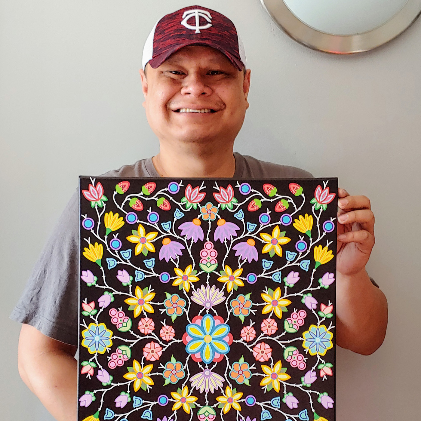 A photograph of Bill Brien wearing a ball cap and smiling while holding one of his paintings based off his Berries, Bee’s and Beauty design. The painting depicts a colorful canvas of yellow, blue and purple flowers, small strawberries and other natural elements in a symmetrical layout over a black background.