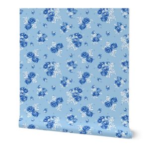 A swatch of wallpaper is placed in front of a white wall. The design on the wallpaper has bright blue cabbage roses, grouped two together, with white leaves, floating amidst a light blue background. Smaller single white and dark blue cabbage roses float in between the rose pairs.