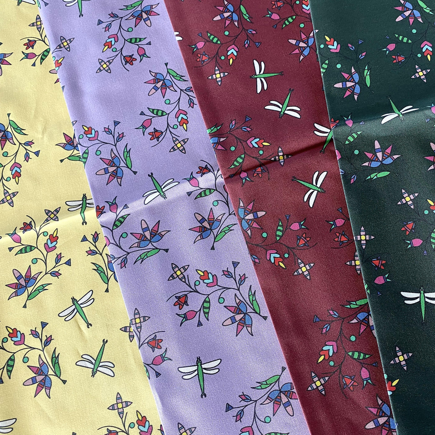 Cole Redhorse Johnson's Dakota Calico design is displayed in four colors, lemon, lavender, maroon and forest green. This repeating design features dragonflies with white wings and green bodies. Small clusters of lavender, blue and maroon flowers growing on green stems with green leaves surround the dragonflies.