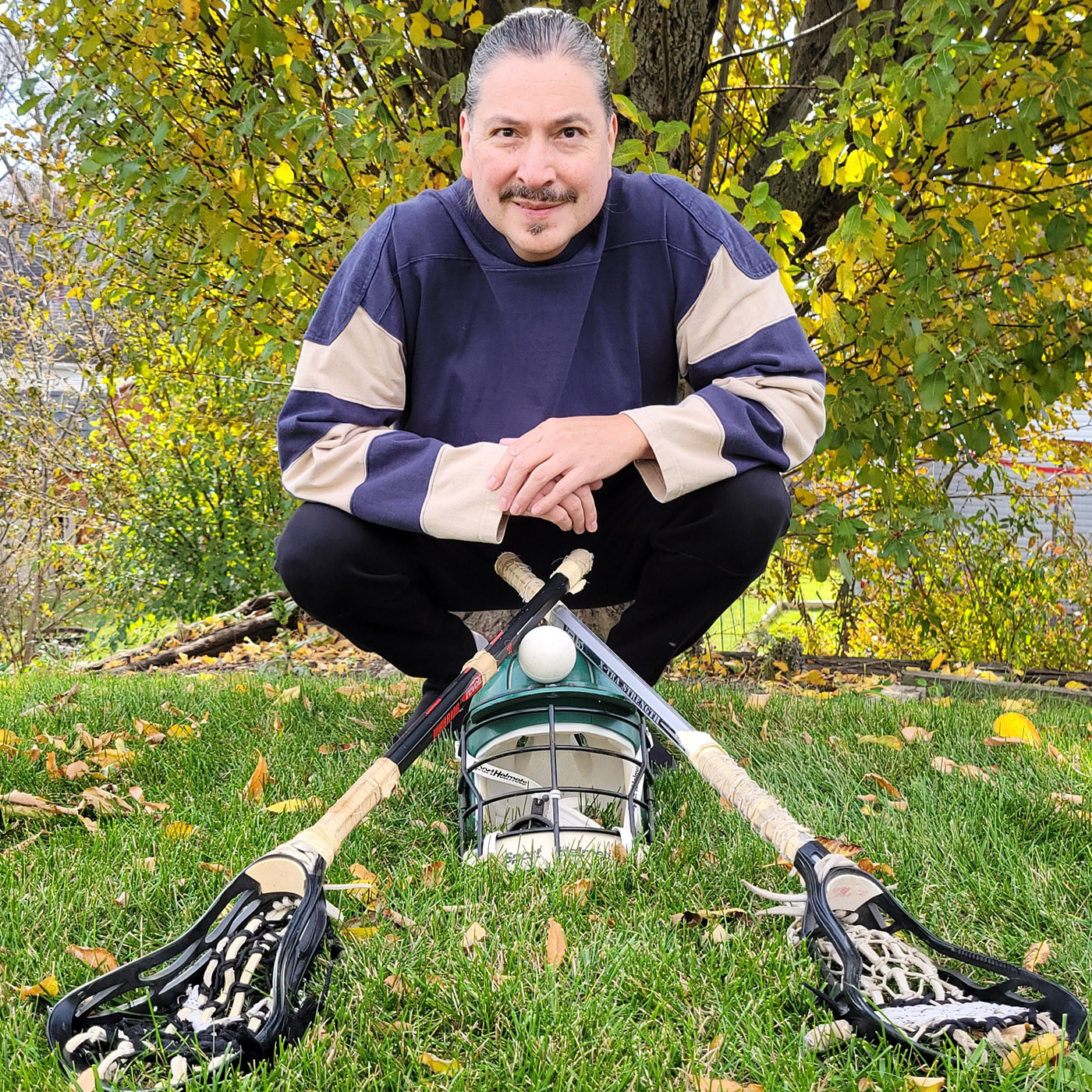 Photograph portrait of Don D Basina smiling and crouched down in the grass on a sunny day in front of a pair of lacrosse sticks that inspired some dd_baz designs.