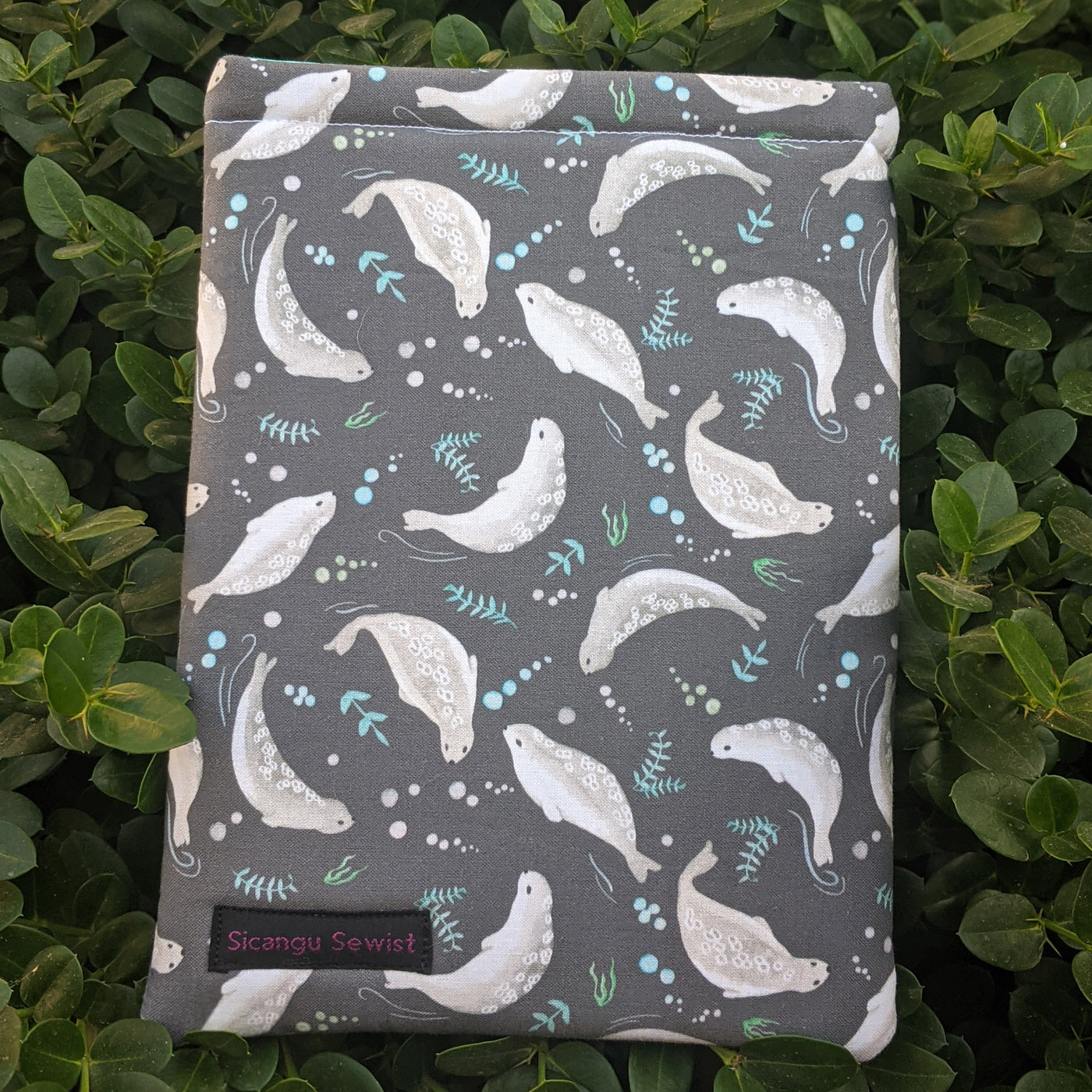 Alt text: A medium-sized handmade book sleeve created by Lorna "Emmy" Her Many Horses out of a fabric that features playful grey seals (or sea lions) and small plant-like shapes in an aqua color. Available on Emmy’s Etsy shop, The Sičángu Sewist for USD$22.
