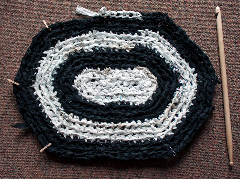 A black and white crocheted rag rug with white fabric in the center, and then black and white circles outside of it in rotating strips of color. A wooden crochet hook lays next to the rug. The hook was fashioned out of a drum stick and the rug is almost as tall as the drumstick/crochet hook. 