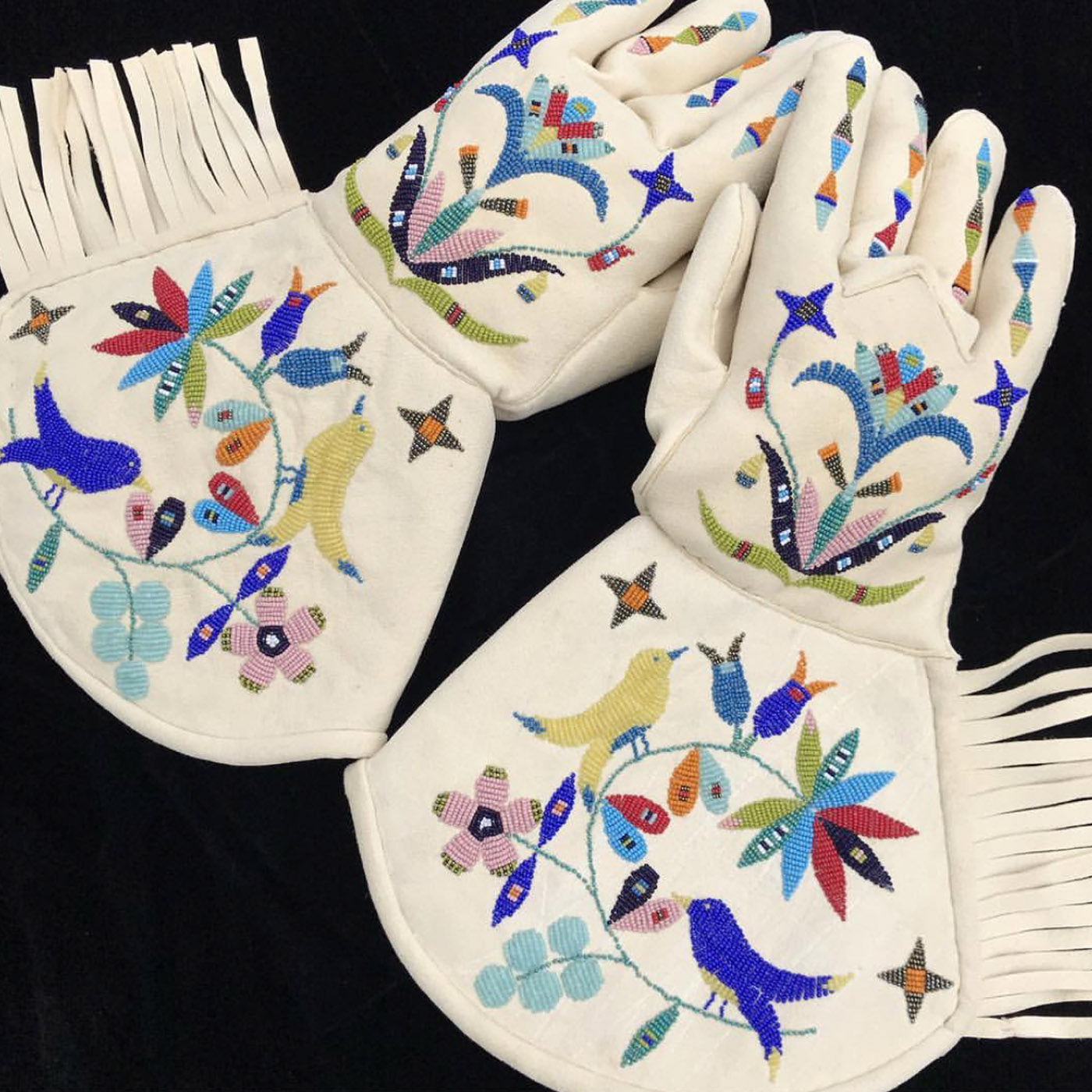 Photograph of a pair of beaded gauntlets handmade by Holly Young. The beaded pattern features an intricate composition of yellow and blue birds standing on an abstract vine of vegetation. The fingers of the gloves hold diamond shaped beadwork. These white gauntlets also feature a fringe.