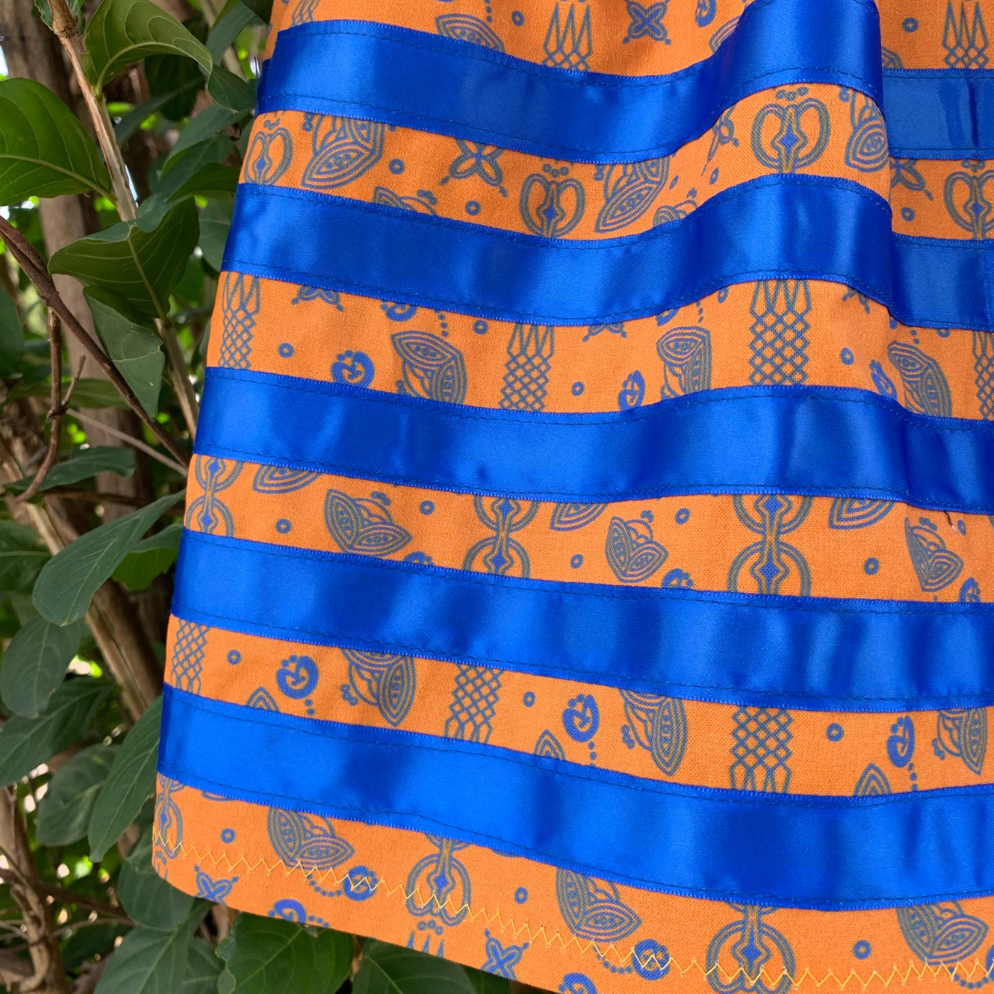 A close up of a ribbon skirt with Jessica's Floral Sunset Gold/Royal design and royal blue ribbons in alternating horizontal stripes. This design has an orange base with small-scale graphic icons in royal blue. The pattern creates vertical lines across the width of the fabric by the way the elements of design are positioned.