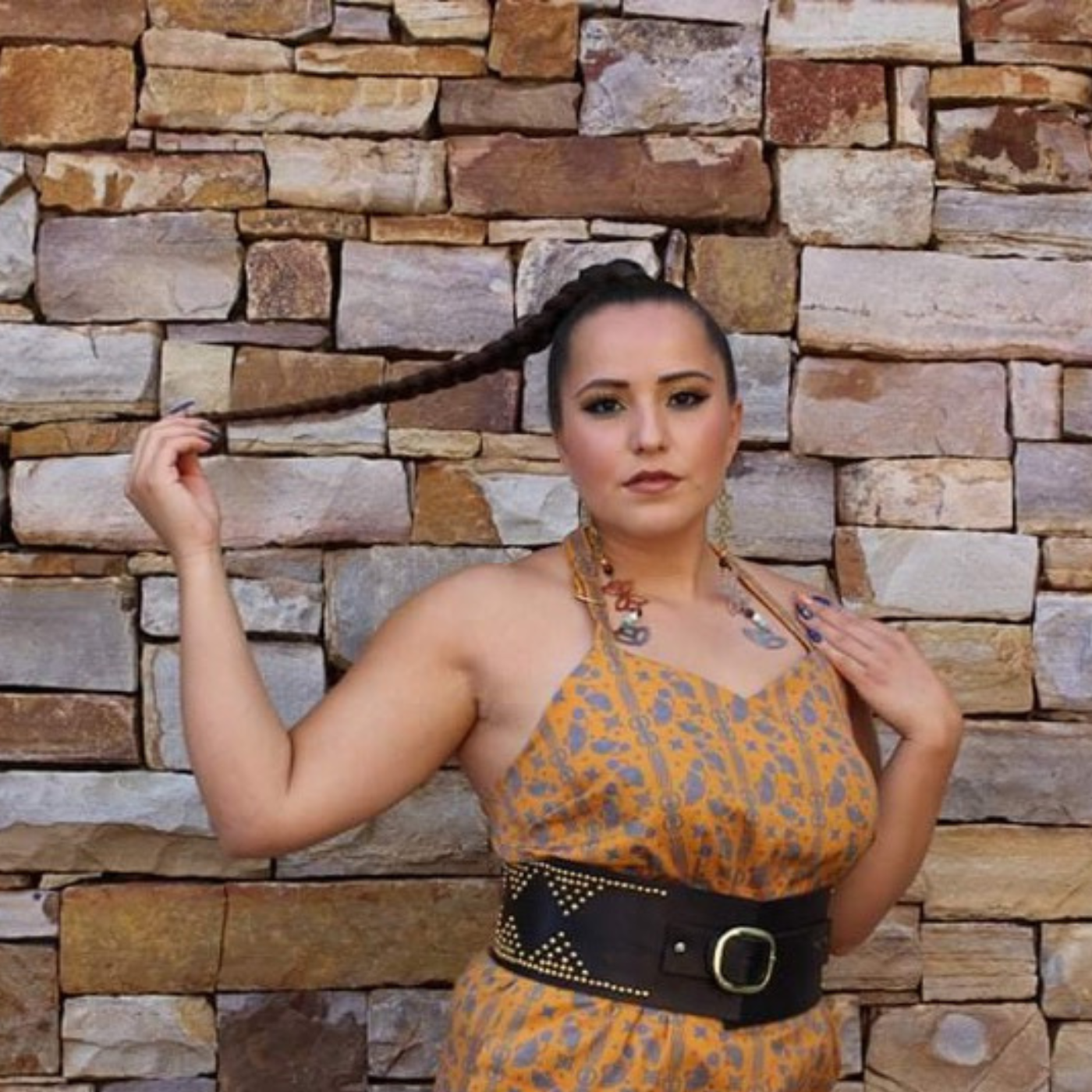 A model wears a jumpsuit with Jessica’s Floral Sunset Gold/Royal design and a white black belt. This design has an orange base with small-scale graphic icons in royal blue. The pattern creates vertical lines across the width of the fabric by the way the elements of design are positioned.