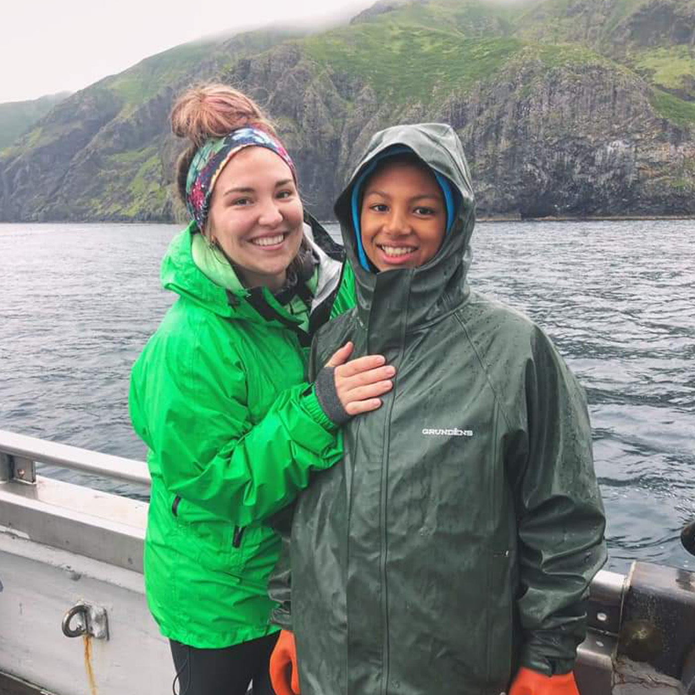 Malerie standing on a boat in Alaska with her family member. They are both smiling despite the cloudy and rainy day. Beautiful mountains can be seen behind them.