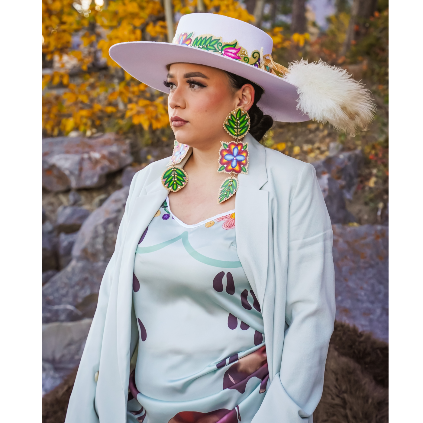 Photograph of Osamuskwasis Roan standing outside during autumn and wearing their handmade clothing: A white hat featuring a band of bright florals and a feather accessory. Osamuskwasis is also wearing oversized earrings from their online shop with the same floral patterns of the har. Finally, a silk dress in soft blue with smaller elements of pink and purple throughout.