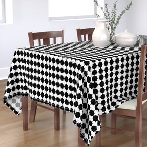 A black-and-white tablecloth lays on top of a dark wooden table with two chairs on one side and the edge of another chair on the other. The design on the white tablecloth is a black grid of squares connected by thin black lines through the centers, both vertically and horizontally. At the points at which the lines meet the squares, there are small half circles.