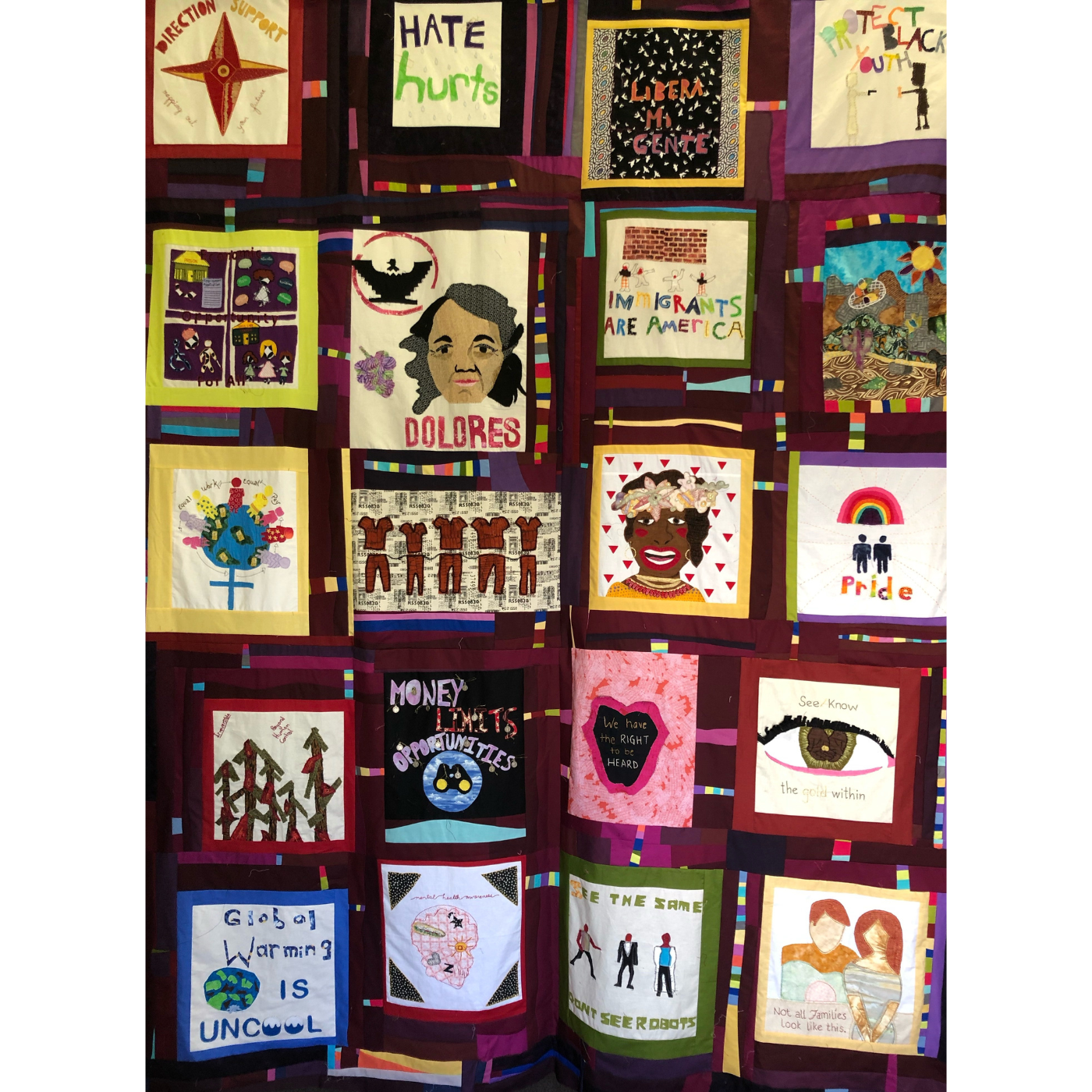 A full size quilt is on display. The 19 blocks in the quilt, which has a burgundy background, cover many social justice issues including global warming, protecting Black youth, gay pride and more.