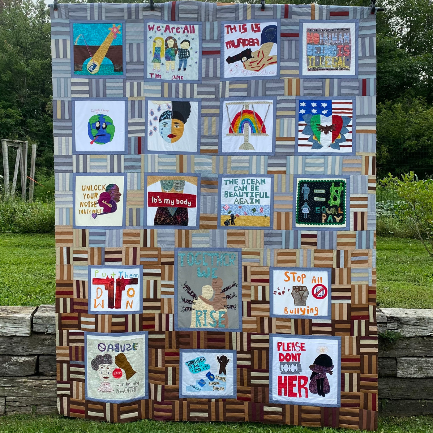 A full size quilt is on display in front a low rock wall with a grassy field behind it. There are 19 quilt blocks on the quilt, which has a gray background at the top and a brown background at the bottom. The blocks in the quilt cover a range of social justice issues from gay rights to ending gun violence to environmental issues to ending genital mutilation and more.