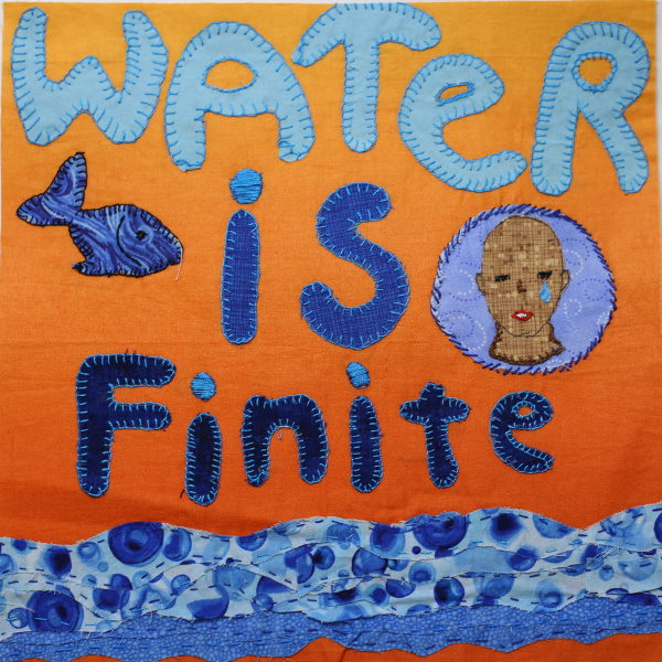 A quilt block with an orange fabric background. The phrase “WATER is finite” is written in blue fabric on the block. The word “water" is in all capital letters and light blue fabric and is at the top of the block. In the middle of the block is the word “is” in medium blue with a blue and white fish to the left of it and the neck and shoulders of a person with brown skin, white teeth and red lips and a blue tear coming out of their left eye to the right. The word “finite” is in dark blue fabric near the bottom of the block. Rippled blue fabric, to represent water, runs across the bottom of the block.