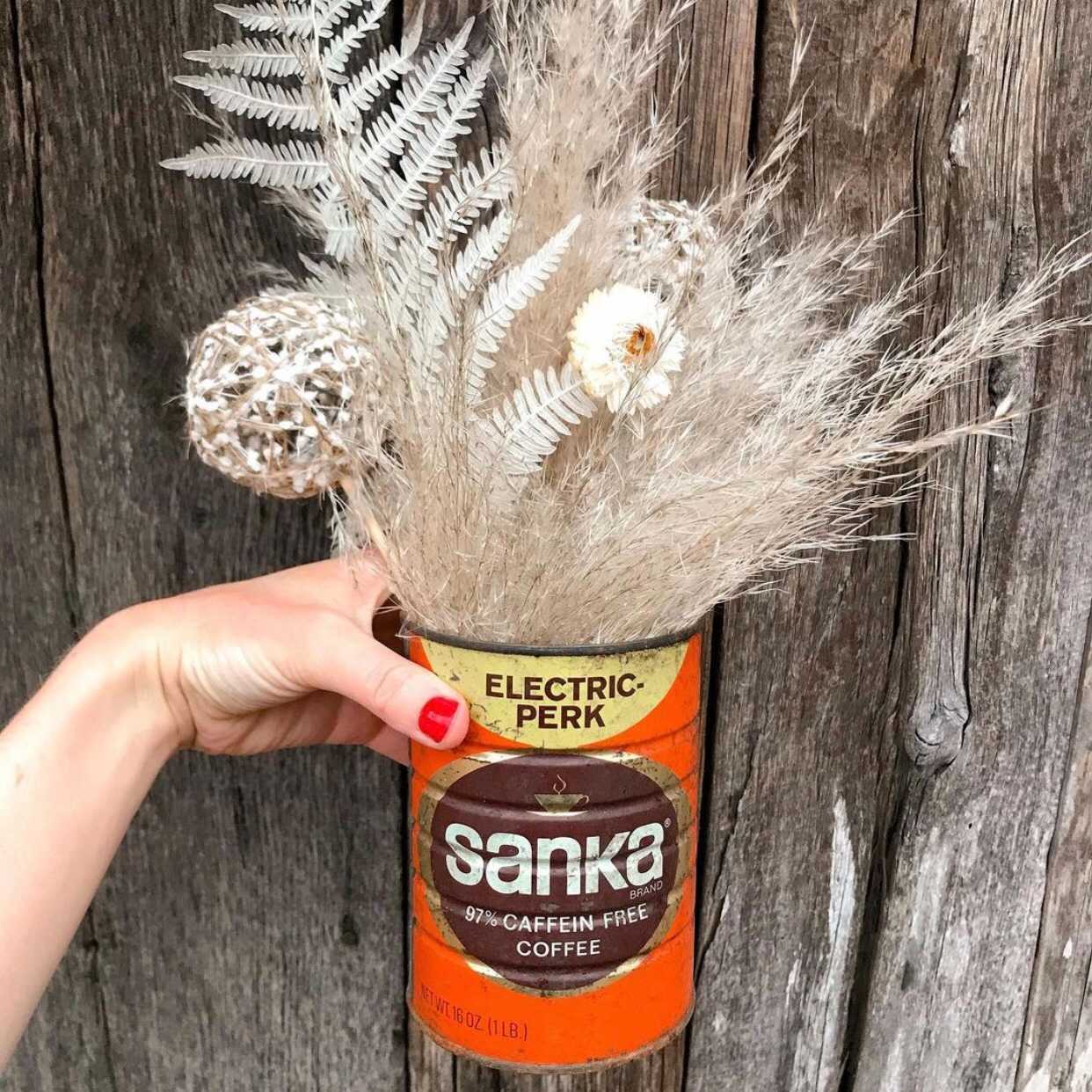 A hand holds a vintage Sanka coffee can filled with beige dried flowers and grasses in front of a gray wooden wall