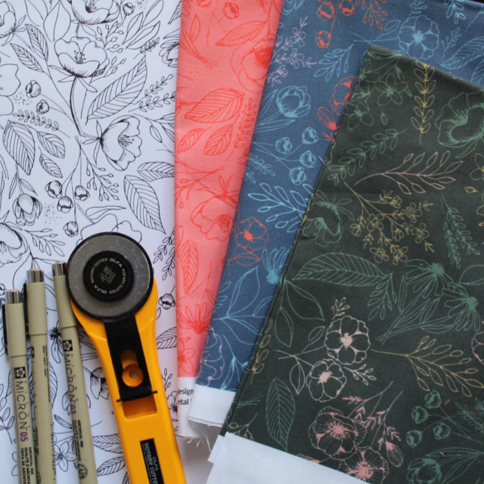 Three folded pieces of fabric, all with a floral design, one red with dark red flowers, one dark blue with light blue, lavender, and red flowers and one black with light pink, dark pink and sage flowers lay next to a white piece of paper filled with a floral design drawn in black. A rotary cutter with an orange handle and three pens lay at the bottom of the piece of paper.