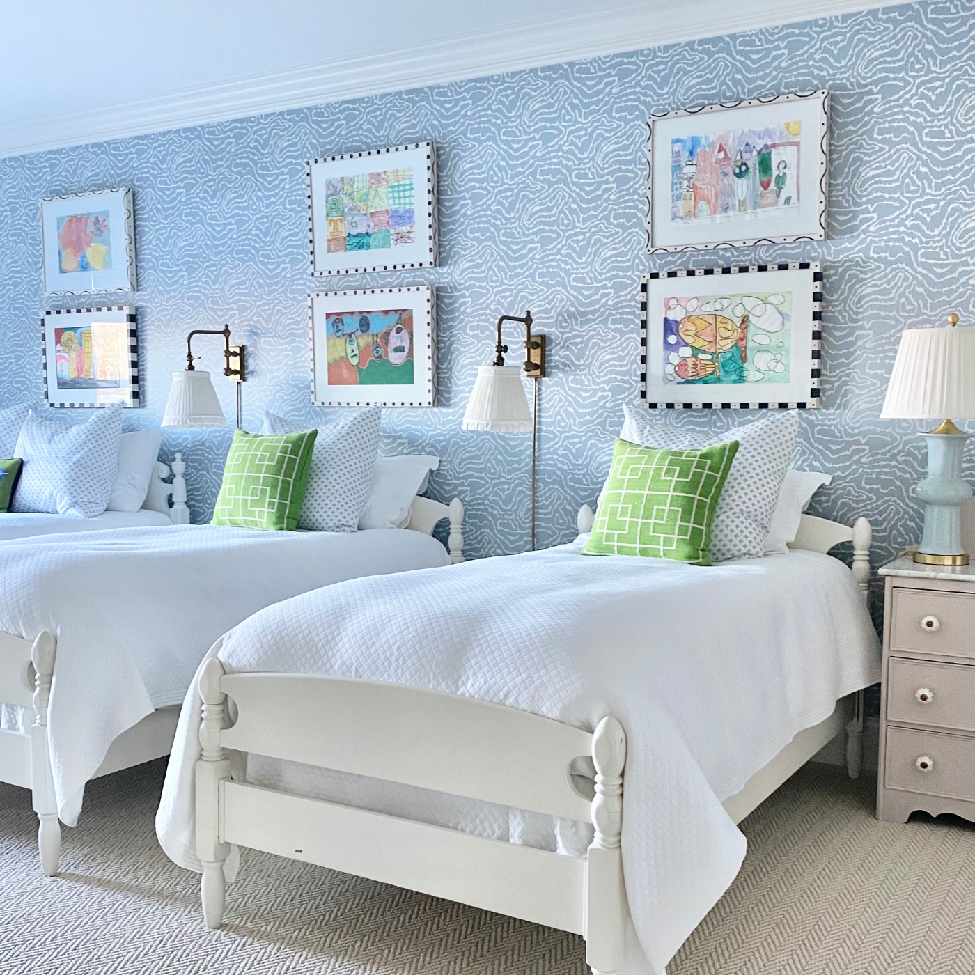Three white twin beds are next to each other in a bedroom with light purple blue wallpaper with white squiggly lines throughout. Two framed colorful prints are above each of the beds. Two white pillows are on each bed behind a green pillow with a white trellis design. Small bronze lamps with white lampshades are on either side of the middle bed. A small white dresser is to the right of the bed in the foreground, it has a light blue lamp on it with a white lampshade.