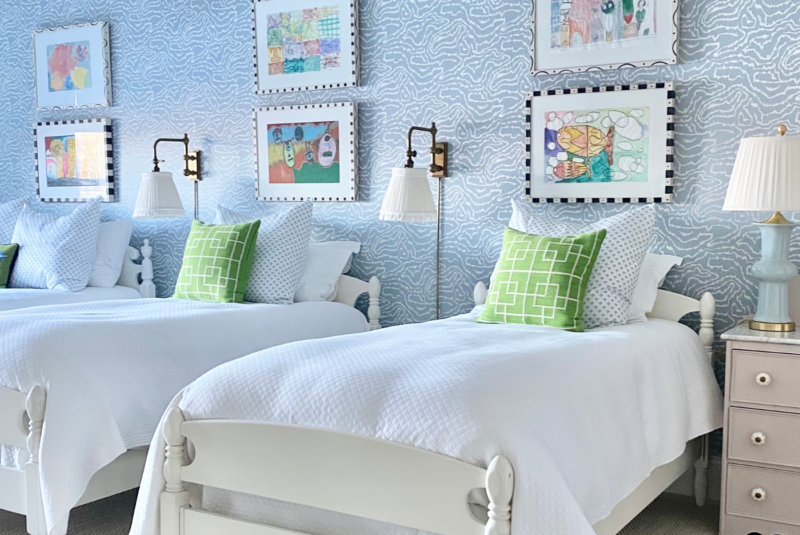 Three white twin beds are next to each other in a bedroom with light purple blue wallpaper with white squiggly lines throughout. Two framed colorful prints are above each of the beds. Two white pillows are on each bed behind a green pillow with a white trellis design. Small bronze lamps with white lampshades are on either side of the middle bed. A small white dresser is to the right of the bed in the foreground, it has a light blue lamp on it with a white lampshade.