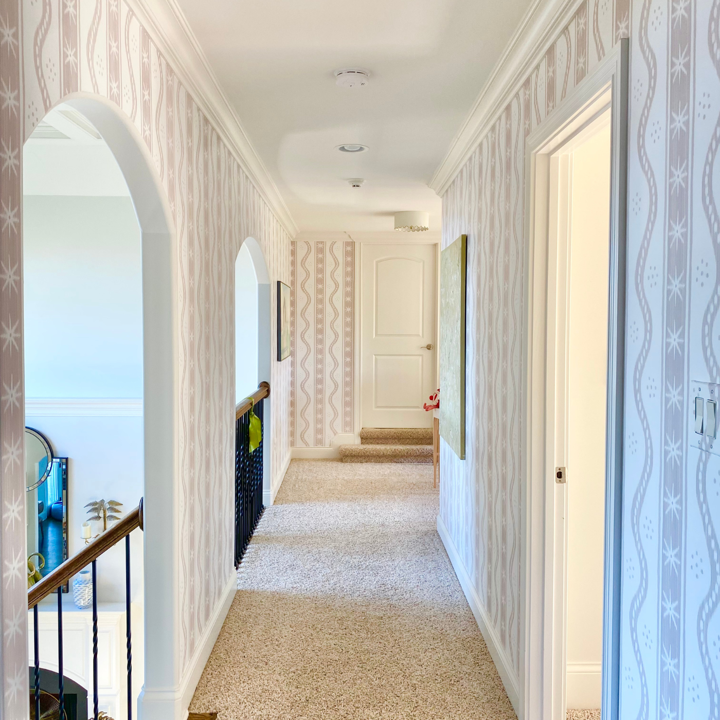 A photo of a hallway with Danika Herrick’s Large Putty on White Charlie Stripe wallpaper. The design has a cream background and columns of white cream stars in long blocks that are light tannish brown alternating with two wavy tannish brown lines close together connected by small tannish brown lines. Small groupings of seven close together tannish brown lines are between the columns with stars and the columns of wavy lines.