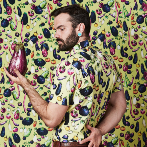 Anthony wears a shirt with a yellow background and purple eggplants, red tomatoes and other vegetables and burgundy pants. He stands in front of a wall with the same eggplant print and is holding an eggplant.