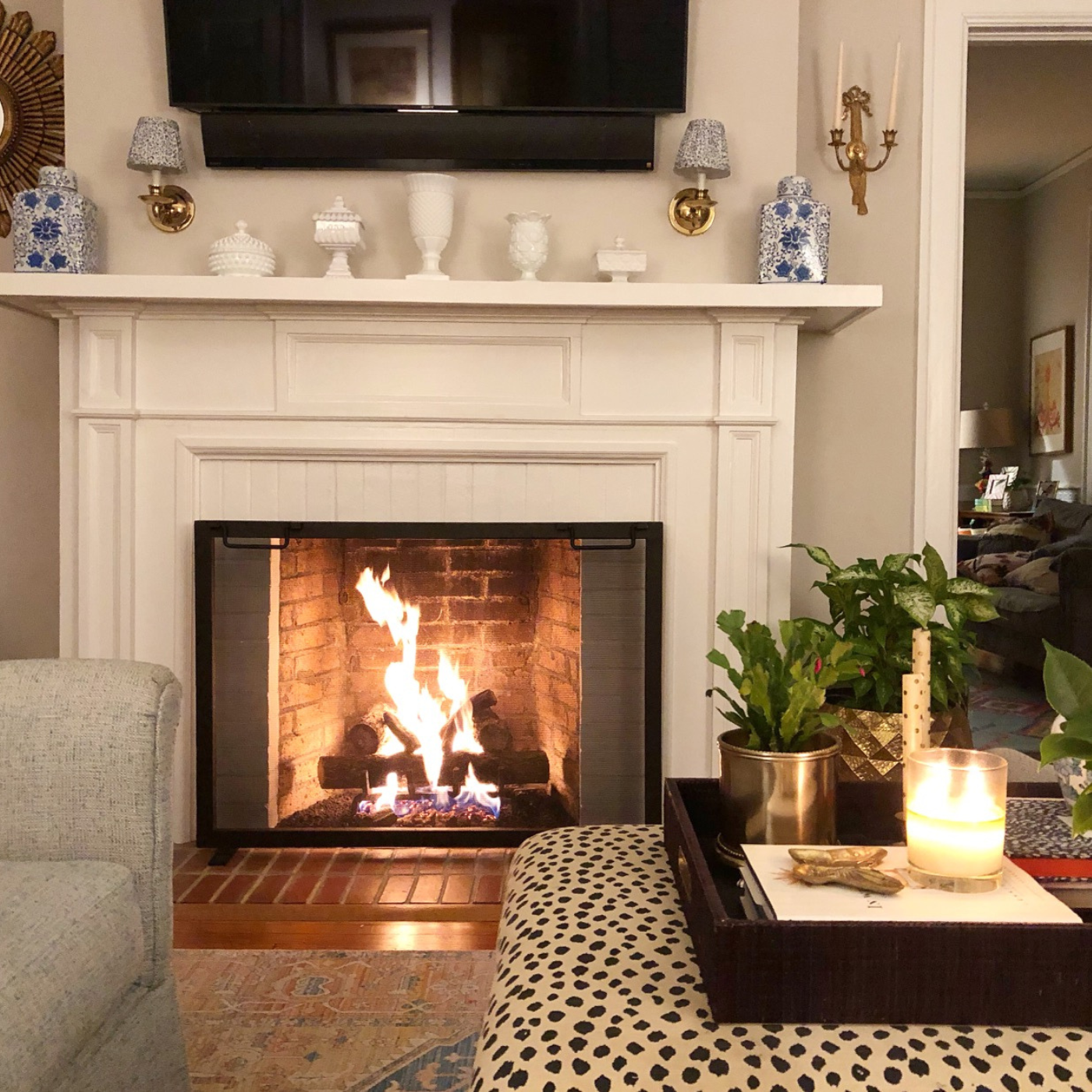 Photo of a living room designed by Cameron. A gray couch is seen partially on the lefthand side of the photo with a roaring fireplace as the focal point. The fireplace has a white mantel with matching blue-and-white vases at either end and small white vases and containers in the middle. Small blue-and-white lamps are above the mantel with a large television screen between them. A white ottoman with black dots is in the right foreground, with plants and a lit candle on it. Another room with a couch is connected to the living room and visible on the righthand side of the photo.