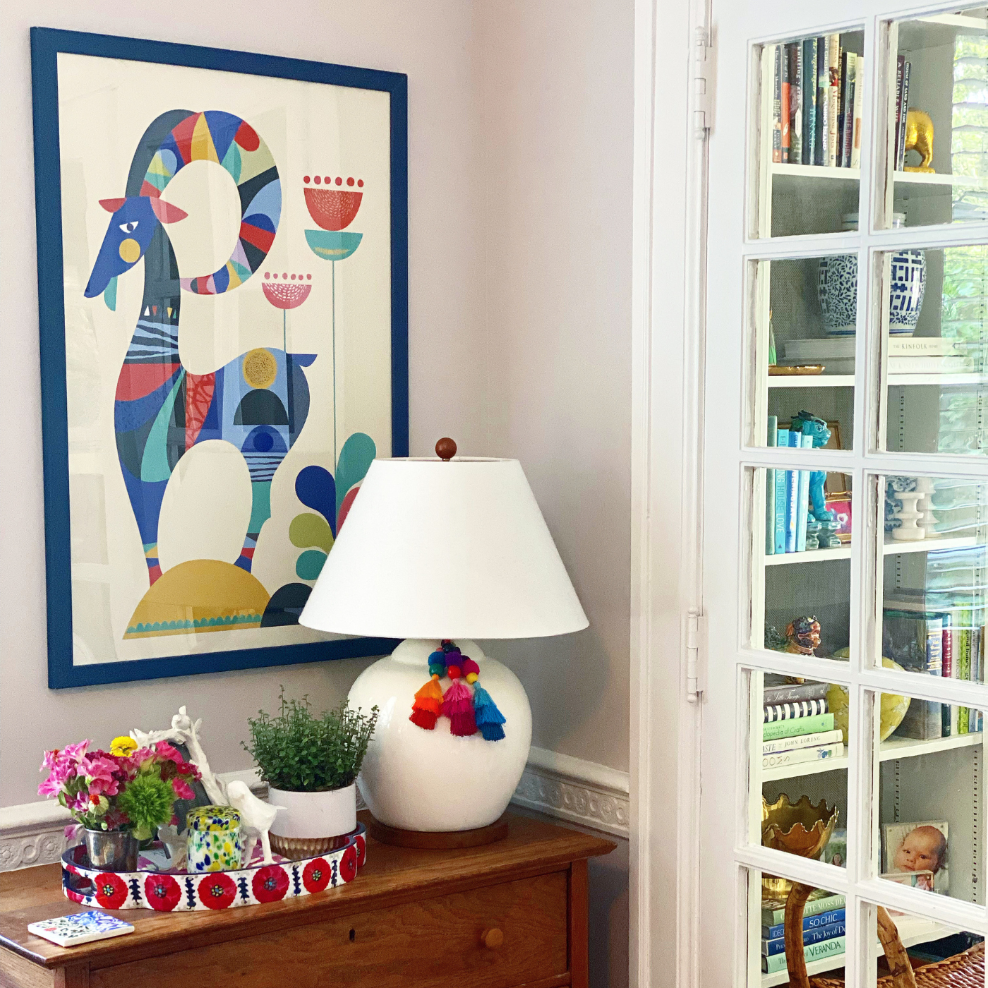 A corner of a playroom, featuring a wooden dresser with a large print of an illustration of a blue goat standing on a small green hill with two red flowers hanging on the wall over the dresser. On the dresser is a small white tray with red floral accents on its edge. On the tray are small flower arrangements. A low white lamp is to the right of the tray. It has a white lampshade and fabric tassels in blue, orange and red. A room with a built-in bookshelf filled with books and small knick knacks is visible through a glass-paned door to the right of the lamp.