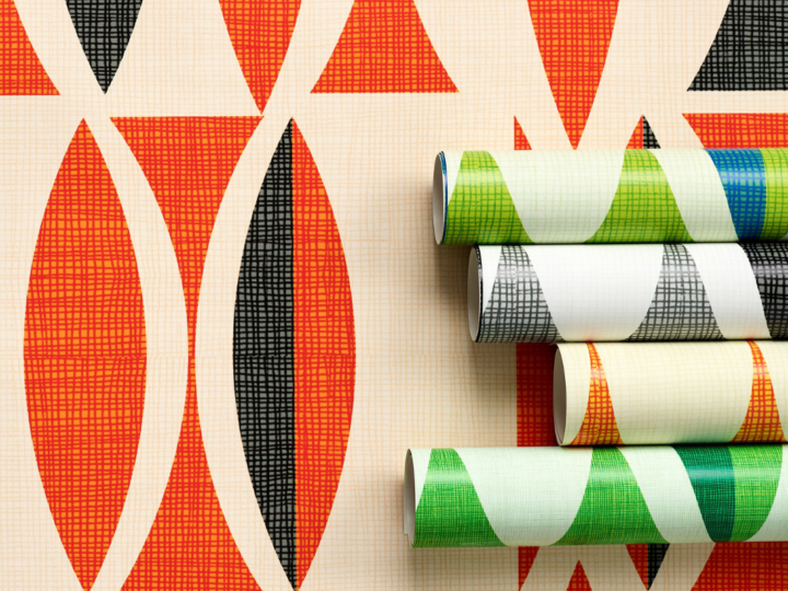 A panel of wallpaper with repeating rows of narrow ovals with pointed tops and bottoms on a cream background is shown laying as if installed on a wall it is taking up the entire background of the image. The ovals are randomly black and dark orange. In the middle third on the right-hand side of the image, four rolls of the same wallpaper design in different colorways are laid lengthwise on top of the black-and-dark-orange panel. Only about a fourth of the rolls are shown. The rolls of wallpaper from top to bottom feature green-and-blue ovals on a cream background, black-and-white ovals on a cream background, orange-and-gray ovals on a cream background and kelly-green-and-dark-green ovals on a cream background.