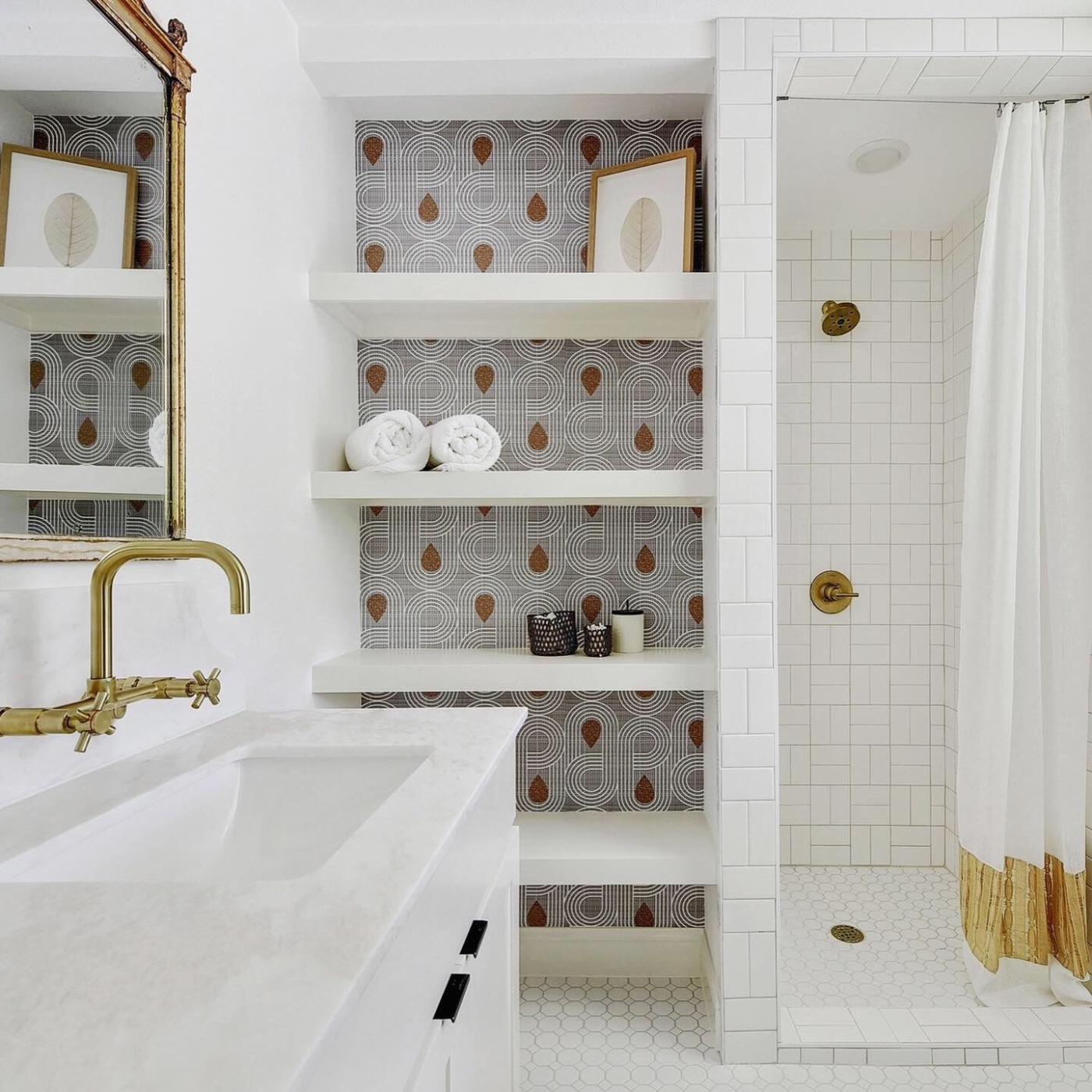 A white bathroom with round white floor tiles and square white bathroom tile has built-in shelves with white round geometric wavy shapes looping in circles on a black background. Small bronze tear drops are in the center of some of the wavy shapes.