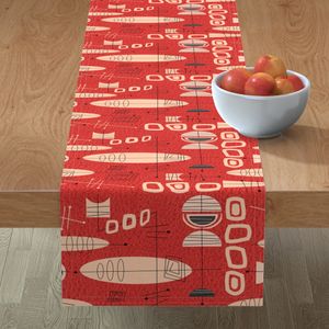 A red and cream mid-century modern and atomic inspired table runner with black accents and ovals, rectangles and an intersecting lines pattern lays across a woodle table with a white bowl of red apples on top of it.