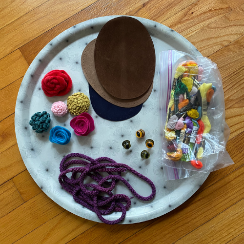 A white round tray with small black starbursts sits on a wooden floor. Several pieces of round brown leather, small fabric flowers, a plastic bag of embroidery thread, glass eyes and purple rope sit on the tray.
