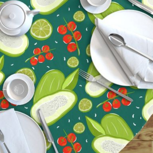A print with green and red vegetables on a dark green background is on a tablecloth partially viewed from above. Two settings of white plates and napkins with silver cutlery are on the table with a white ceramic coffee pot and coffee cup.