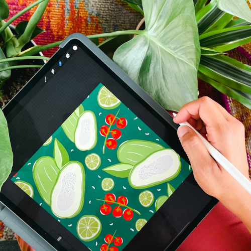 Stephanie sits with a tablet on her lap with a drawing of green and red vegetables on a dark green background on it, she is surrounded by plants. A digital pen is in her hand, which sits on the right edge of the tablet.