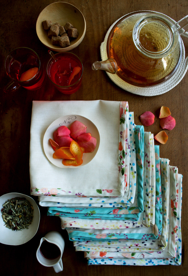 A small white saucer full of red and orange flower petals sits on a stack of white napkins, each with a different fabric edge, some white floral, some blue floral, some blue polka dot. A glass tea pot, a small brown bowl of brown sugar, two glasses are to the top and right of the fabric. A small bowl of loose green tea and a small white ceramic pitcher with brown liquid are on the bottom left-hand side of the fabric. 