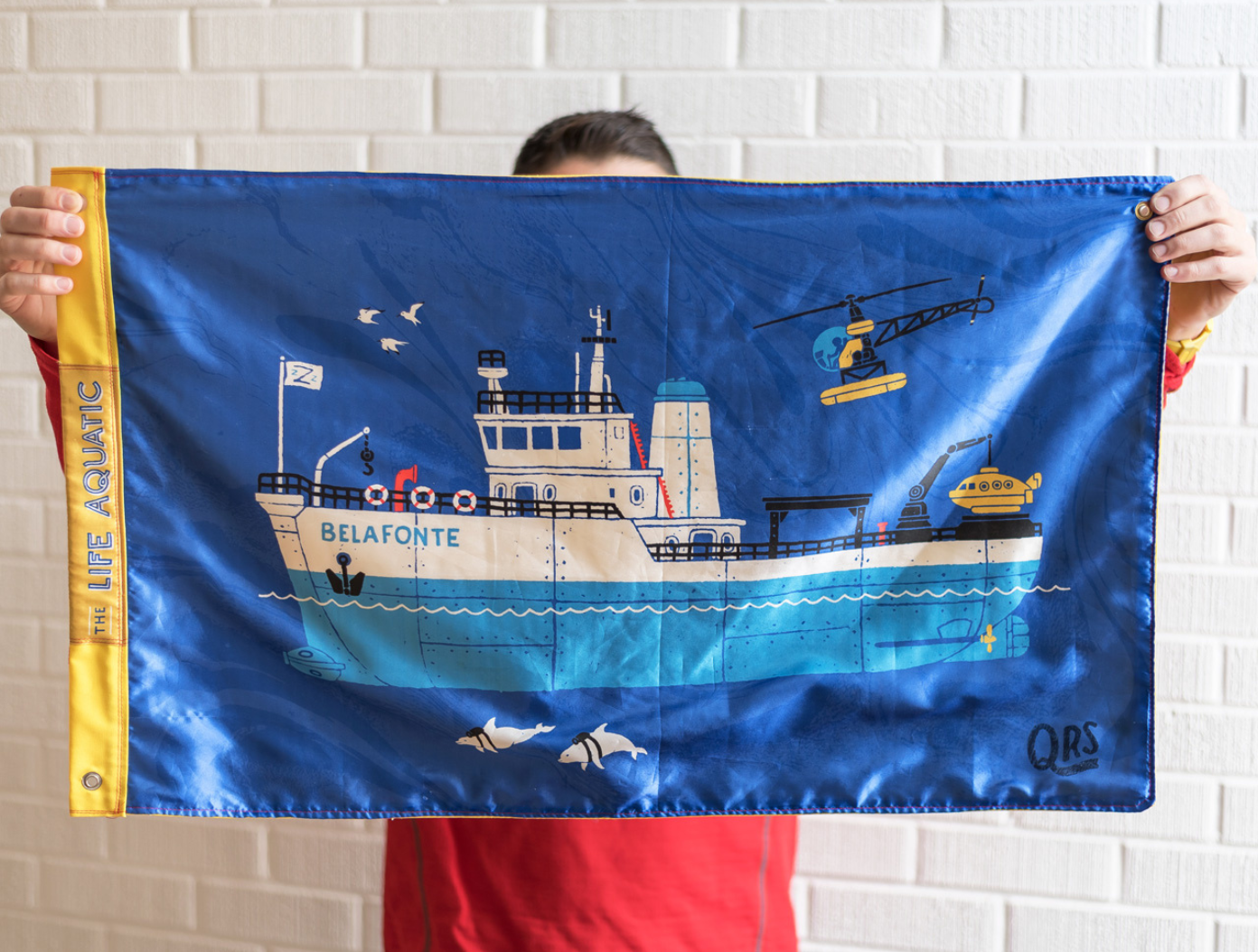 Two hands hold up a flag with a drawing of the Belafonte, the ship in the movie The Life Aquatic. Along the left edge of the flag is a yellow strip that has the movie title in all white capital letters. The ship is turquoise on the bottom half and white on the top half. A small yellow underwater research vessel is at the back of the ship and a yellow-and-black helicopter flies overhead. The front of the ship has white flag with a blue turquoise letter “Z” on it and the word “Belafonte” is in turquoise at the front of the ship.