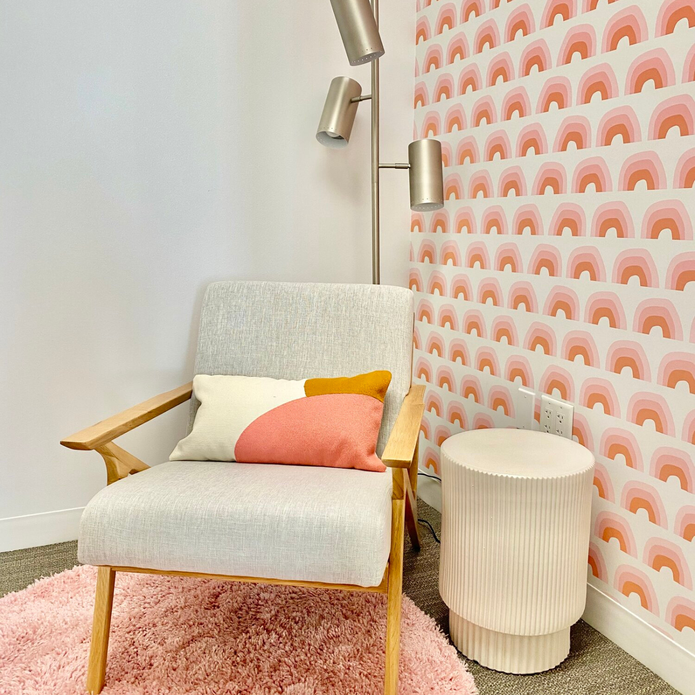 A gray fabric chair with thin wooden arms and legs and a white, brown and pink pillow on top sits on a small round pink shag rug. A silver lamp with three light bulbs sits behind it and a small round cream table sits to the chair’s left. The wall on the left is cream. The wall on the right has a cream background and repeating rows of small brown and pink rainbows.
