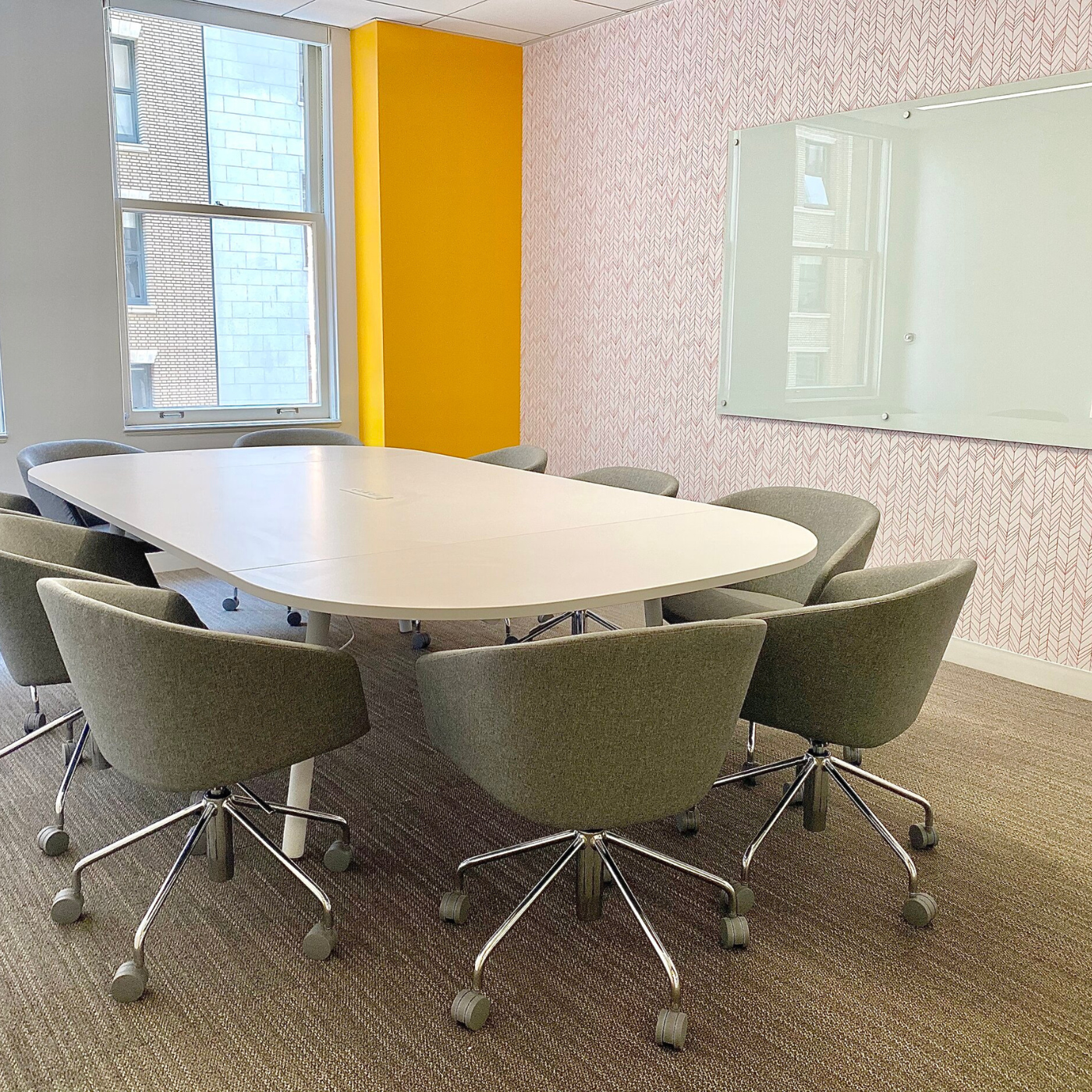 A white oval conference table with 10 gray rolling chairs around it sits in the middle of a room in front of two windows with a brick office building outside. The wall to the right of the windows has been painted dark yellow. On the wall to the right of the table and chairs is a whiteboard on a wall with wallpaper featuring rows of light pink and white herringbone.