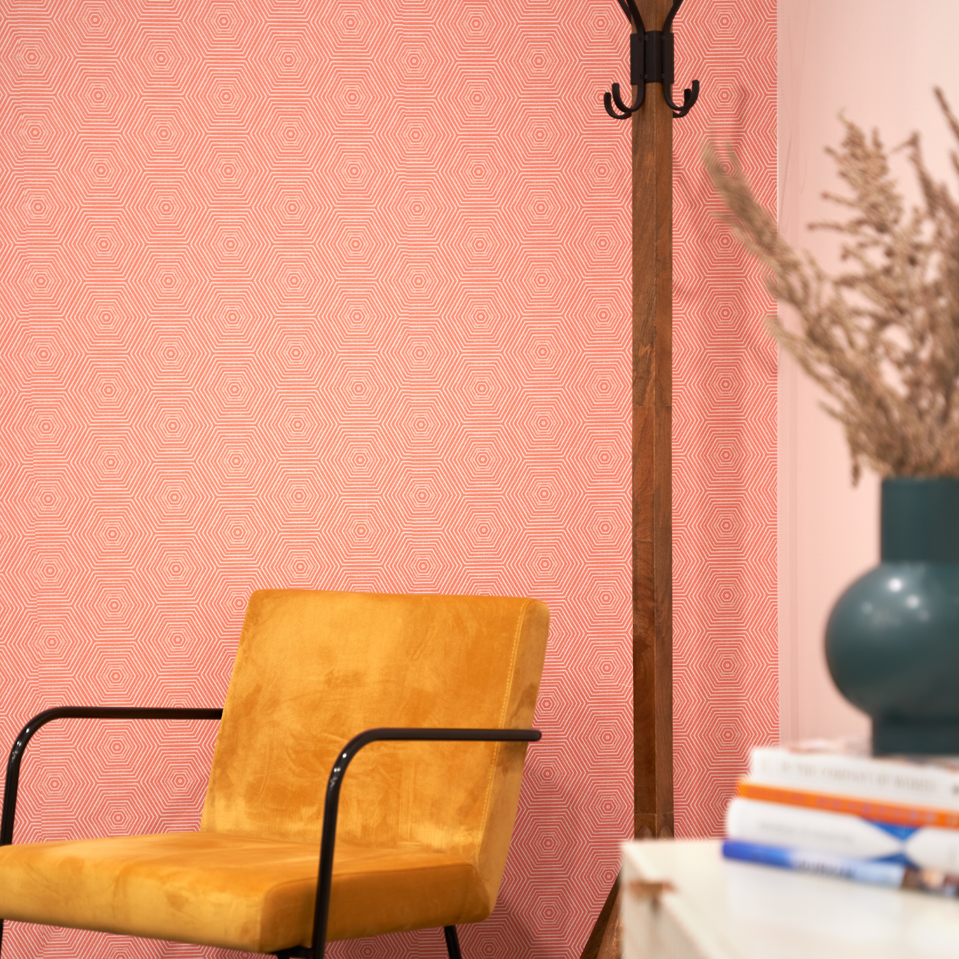A yellow velvet chair sits in the corner in the apex of two walls with pink wallpaper that has a design featuring rows of interconnected small white geometric shapes. Behind the chair is a wooden coat rack and along the wall to the right is a white bookshelf with a dark blue planter on top and dried flowers coming out of it on it as well as a stack of books.