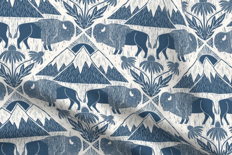 Navy blue linocut print of rows of navy buffalos in pairs of two, facing each other with small triangle designs appearing between the two paired buffalo heads. Underneath the rows of paired buffalos are rows with a repeating design of three navy mountains grouped together with snowy cream tops and a sun with ray peaking out over top the mountains and three wildflowers coming out of leaves.