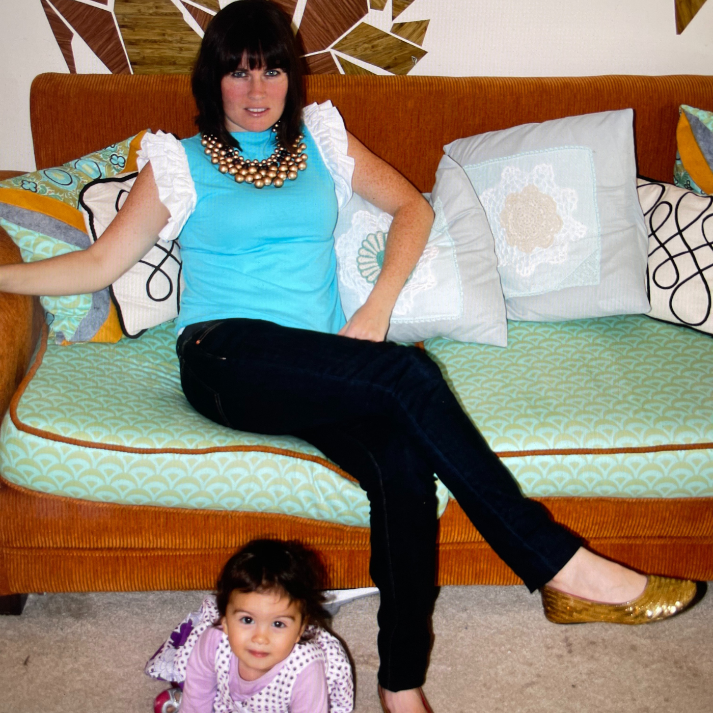 Katie Kortman sits on a mint green couch with a repeated tan half moon design and dark brown piping. She is wearing black pants, brown shoes, a turquoise top with short white ruffled sleeves and a bronze statement necklace consisting of several rows of small orbs linked together. Along the back of the couch are loads of pillows Katie made, some are gray with white doilies, some are white with black squiggles, some are bright green and floral. Behind Katie is a dark brown and light brown mural of geometric shapes on a white wall. Katie’s oldest daughter, when she was a baby, is on the floor in front of her, wearing a purple-and-white outfit. The shirt, couch covering, pillows and mural were all made by Katie.