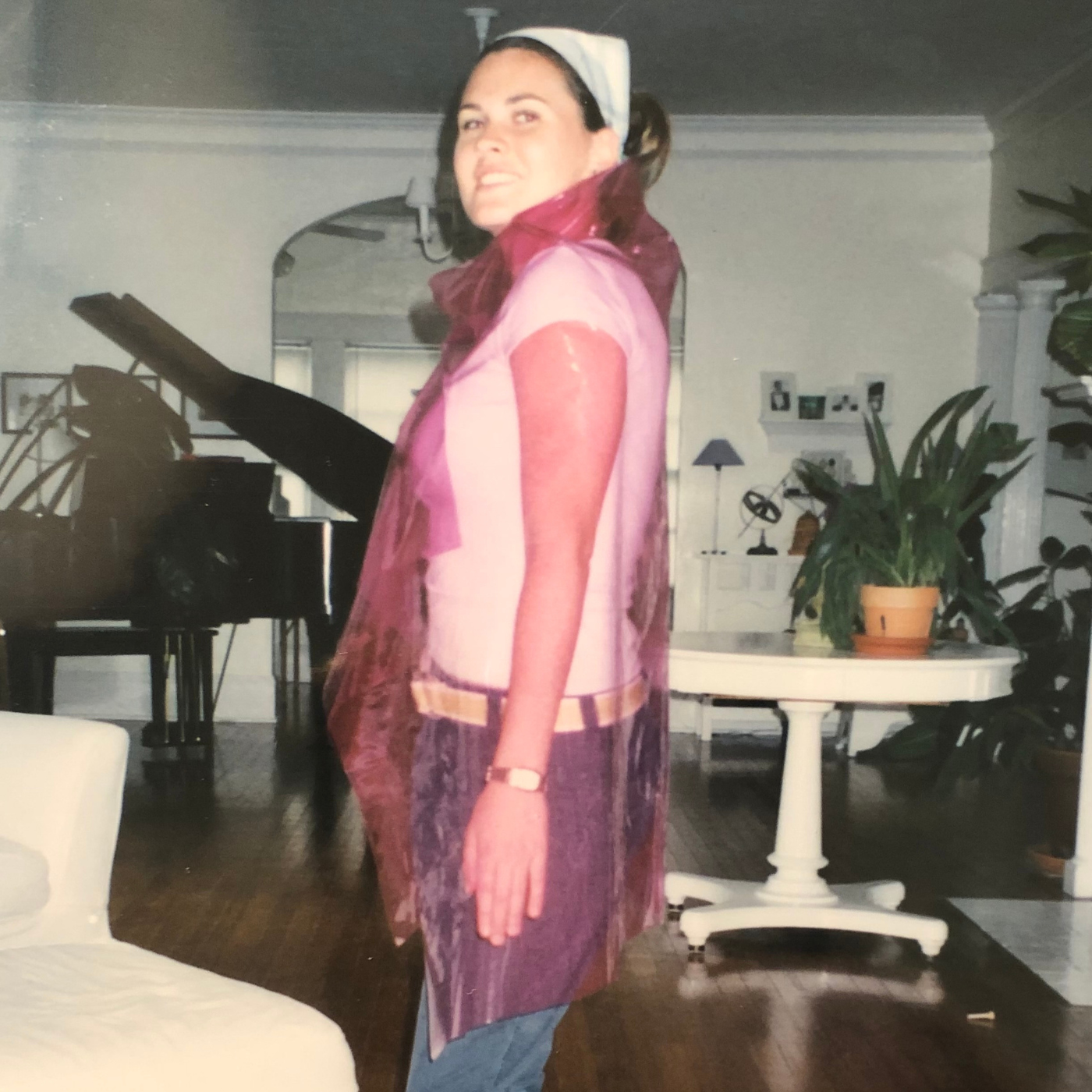 Katie Kortman stands to the side wearing a clear purple rain jacket that goes down her thighs and has a high neck. A white short-sleeved shirt, cream belt and blue pants can be seen underneath. Katie is wearing a white bandana in her hair and standing in a living room, with a black piano to her left and a white table with a potted plant on it to her right.