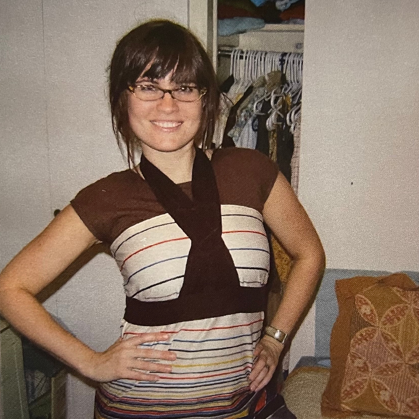 Katie Kortman stands with her hands on her hips wearing a dress made from a thrifted skirt. The dress has brown fabric at the shoulders and then moves into a striped design in cream, yellow, black, red and blue. A strip of dark brown fabric wraps around her chest and around her neck.