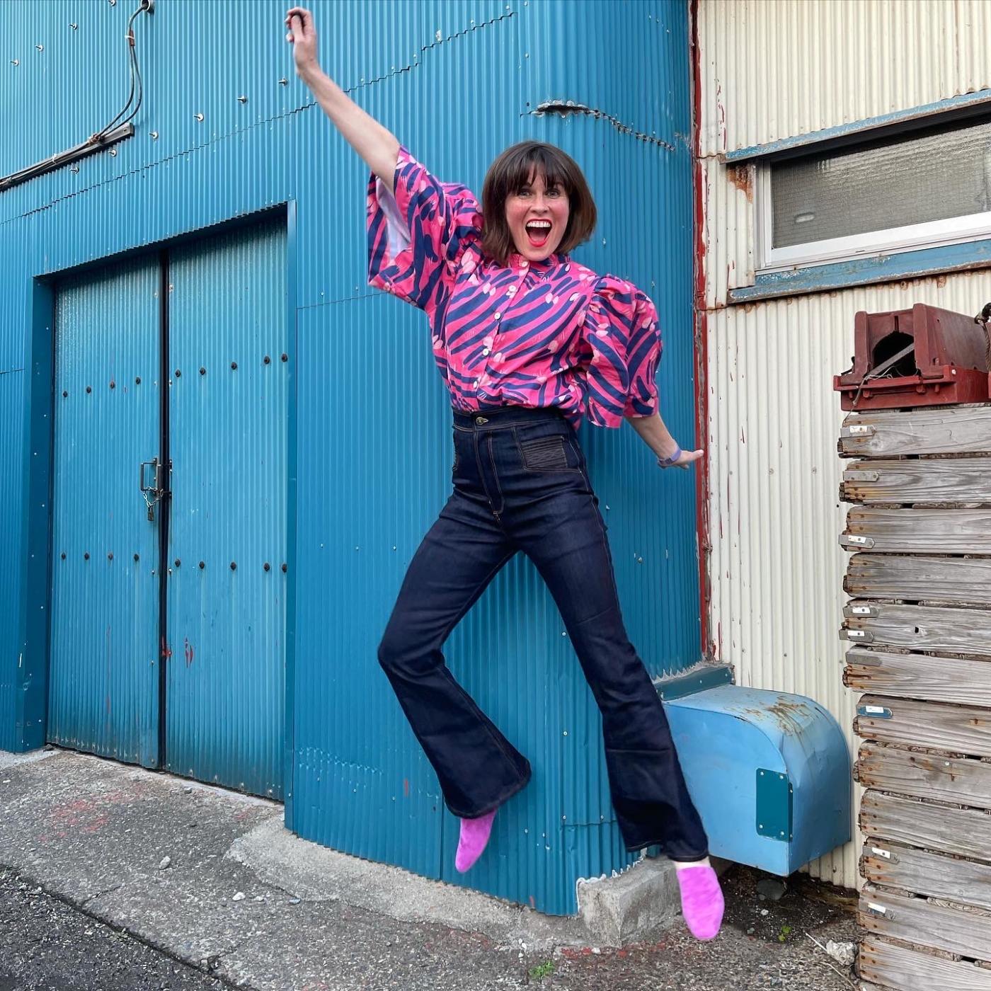 Katie is jumping in the air, with her right arm extended to the right and upward and her left arm facing left and downward. She is wearing a button-up shirt with billowy short sleeves that has a hot pink background and short purple stripes connected by white dots. The pants are high-waisted jeans. Her shoes are hot pink pumps and she is standing at the edge of a bright blue industrial building, where it meets a cream industrial building.