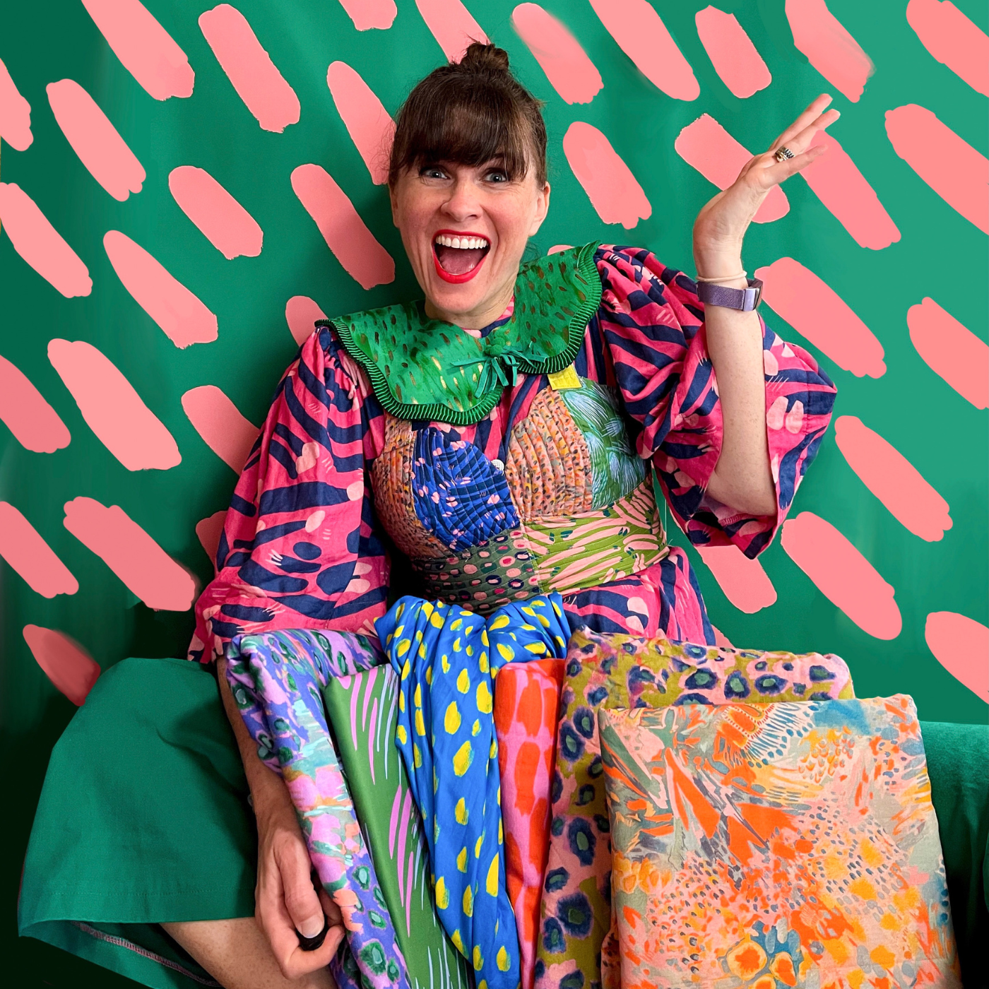 Katie Kortman sits against a green wall with short salmon-colored lines in repeated rows behind her. Her left hand is up in the air and her right hand is down in her lap holding a fabric stack of Katie’s designs that spills toward the camera. The designs are a range of florals and solid backgrounds with short repeated lines in contrasting colors. Katie wears a dress made out of several of her brightly colored floral fabrics with a green detachable collar.