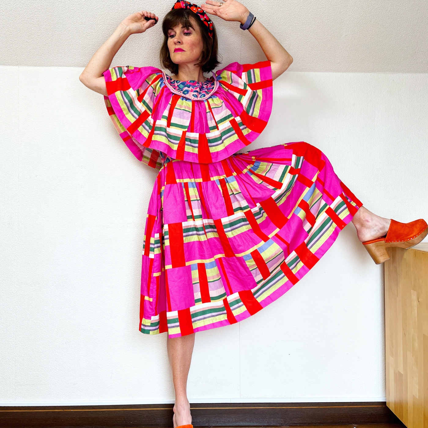 Katie Kortman stands with both hands reaching above her head and her right leg up on a piece of wooden furniture. The dress is knee length and the top has a large section of drapey fabric that looks almost like a cape. The pattern has rows of hot pink and red vertical blocks interspersed by horizontal stripes in pink, blue, white, yellow and green.