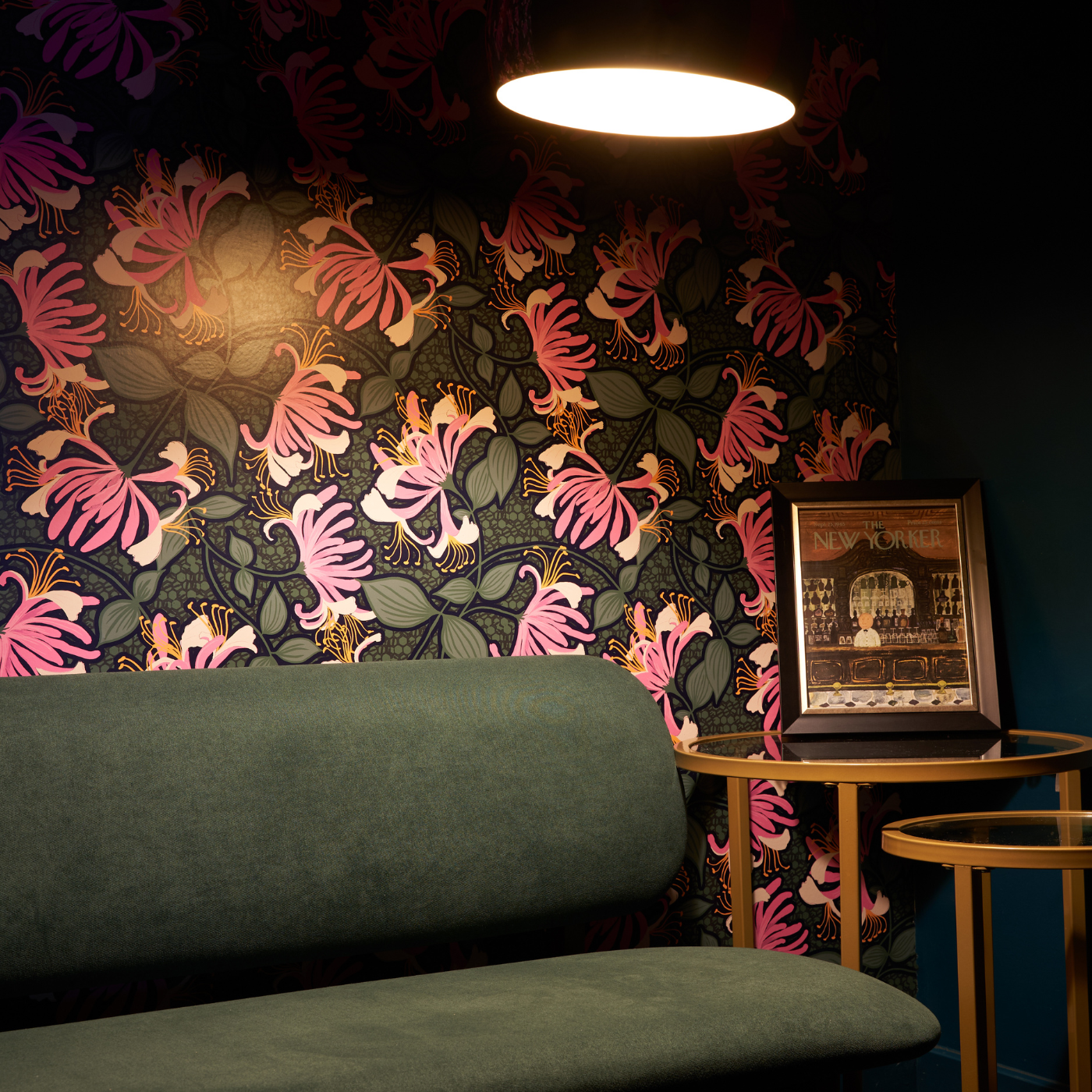 A dark green sofa sits underneath a large tall lamp with a small wooden table beside it. The wallpaper behind the sofa has a black background and hot pink flowers extending in all directions each with a grouping of small green leaves.