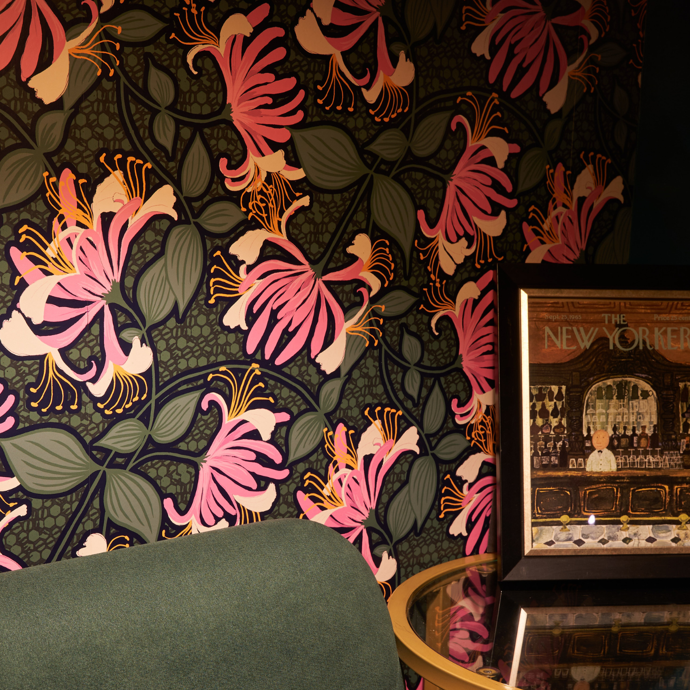 The corner of a dark green sofa in a small room with a small wooden table beside it. The wallpaper behind the sofa has a black background and hot pink flowers extending in all directions each with a grouping of small green leaves.