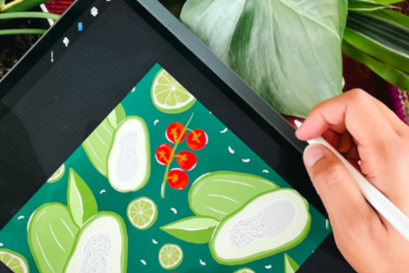Stephanie sits with a tablet on her lap with a drawing of green and red vegetables on a dark green background on it, she is surrounded by plants. A digital pen is in her hand, which sits on the right edge of the tablet.